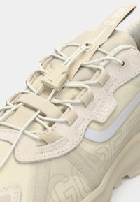 Beżowe buty sportowe z teksturowaną cholewką, elastycznymi sznurówkami i formowanym plastikowym zapięciem w postaci toggle. Zawiera detal z logo "Vans".