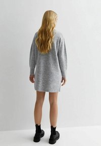 Femme blonde aux cheveux ondulés portant une longue robe-pull grise en maille et des bottines noires, debout devant un mur blanc uni.