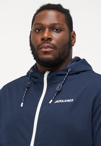 Chaqueta con capucha de color azul marino con un zipper blanco, con detalles de cuerda ajustable. La marca "JACK&JONES" está impresa en blanco en el pecho.