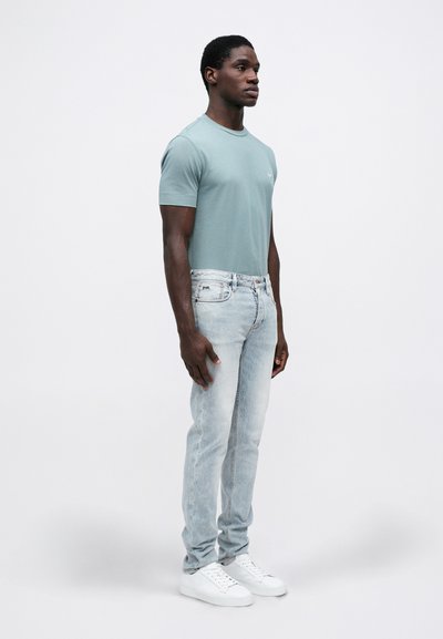 Mann iført en lys blå tettsittende t-skjorte, lyse vaskede skinny jeans og hvite joggesko som står mot en enkel hvit bakgrunn.