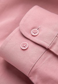 Gros plan sur un poignet de chemise rose avec deux boutons ronds assortis de couleur rose et des coutures détaillées sur un tissu lisse.