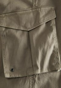 Tkanina khaki z dużą kieszenią na klapie oraz wzmocnieniem w postaci oczek. Gładka tekstura, solidne wzory ściegów i prosty, funkcjonalny design.