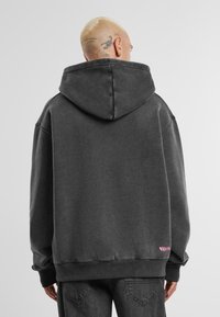 Sötét szürke kapucnis felső, nagy kapucnival, lejjebb eső vállakkal és egy apró rózsaszín logóval az alján. Az anyag lágy és texturált.