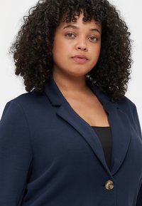 Blazer bleu marine en tissu doux, doté d'un unique bouton en bronze, de revers crantés et d'une coupe décontractée, porté sur un haut noir.