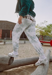 Hedvábně modrý kabát, bílé tepláky s tie-dye vzorem a bílé tenisky se stříbrnými detaily. Stojí na kládě v písčité oblasti na hřišti.