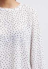 Blouse blanche en tissu léger et texturé avec un motif à pois noirs. Encolure ronde avec un détail délicat de volants.