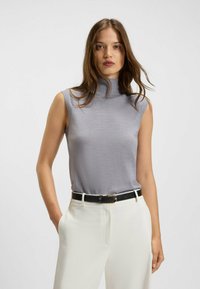 Top senza maniche con collo alto in tessuto grigio chiaro, caratterizzato da una texture liscia. Abbinato a pantaloni bianchi e una cintura nera con una fibbia dorata.