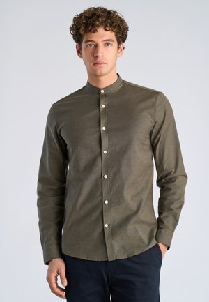 Jonge man met krullend haar, gekleed in een olijfgroen overhemd met mandarin kraag en knopen, en donkere broek, tegen een effen achtergrond.