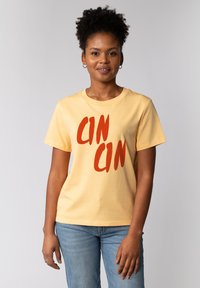 Korteboezen gele katoenen t-shirt met opvallende rode "CIN CIN" tekst in een penseelstreeklettertype, gecombineerd met lichtblauwe spijkerbroek.