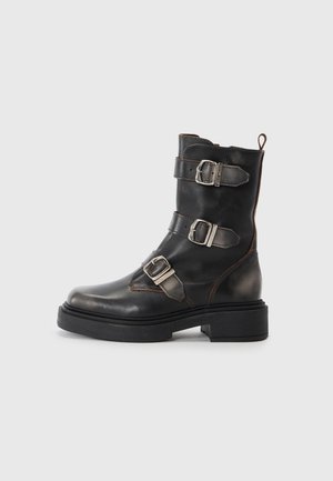 Black leather ankle boot with three metallic buckles, round toe, matte finish, and a thick rubber sole. Features a pull tab at the back.