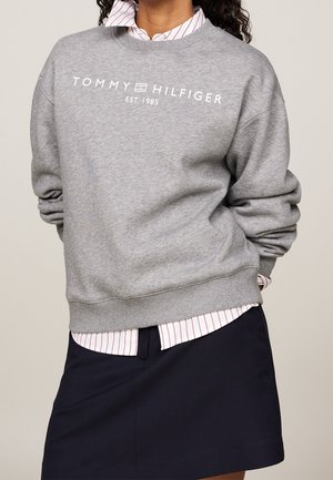 Persoon draagt een grijze Tommy Hilfiger sweater over een gestreept shirt, gecombineerd met een zwarte rok, tegen een effen achtergrond.