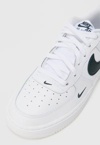 Bijele kožne Nike Air Force 1 tenisice s reljefnim tamnozelenim Swoosh-om, perforiranim prednjim dijelom i bijelim vezicama, koji imaju podstavljeni okovratnik.