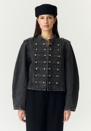 Donna che indossa una giacca di jeans nera stile militare con dettagli a bottoni e un cappello tondo nero, in piedi davanti a uno sfondo uniforme.