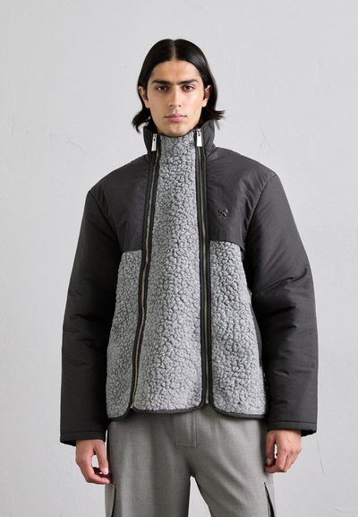 Padded Jacke mit einer schwarzen Außenfläche und einem weichen, grauen Fleece-Innenfutter, hohem Kragen und doppelt ausgeführten Reißverschlüssen.