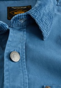 Blå kordfløjlskrave på en skjorte med en glat stofkrop. Har en sølvknap med præget tekst, der fremhæver premium kvalitet.