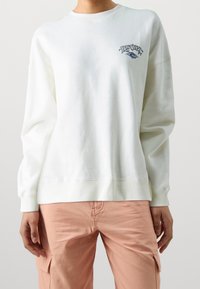 Biała bawełniana bluza z okrągłym dekoltem, długimi rękawami i małym niebieskim logo graficznym na piersi. W połączeniu z różowymi spodniami cargo.