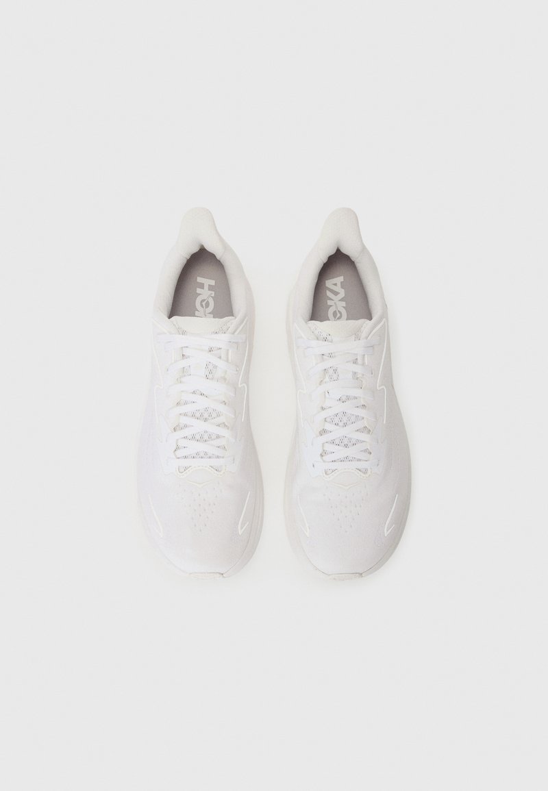 White athletic shoes with a textured upper, reinforced toe, and contrasting gray logo on the insole. Features flat white laces and cushioning.