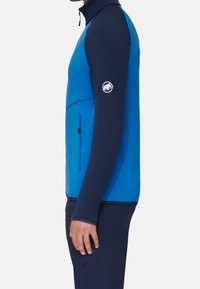Blauwe, getailleerde zip-up jas met hoge kraag en een met rits afsluitbare zak. Heeft contrasterende marineblauwe mouwen en een logo patch op de arm.