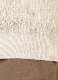 Pull en crème, tricoté avec un tissage texturé et un ourlet côtelé, superposé sur un tissu marron. Présente une construction légère et ample.