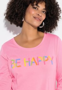 Vaaleanpunainen pitkähihaisen paita, jossa on teksti "OLE ILOINEN" monivärisillä sahalaitakirjaimilla. Kankaan näyttää olevan pehmeä ja siinä on pyöreä kaula-aukko.