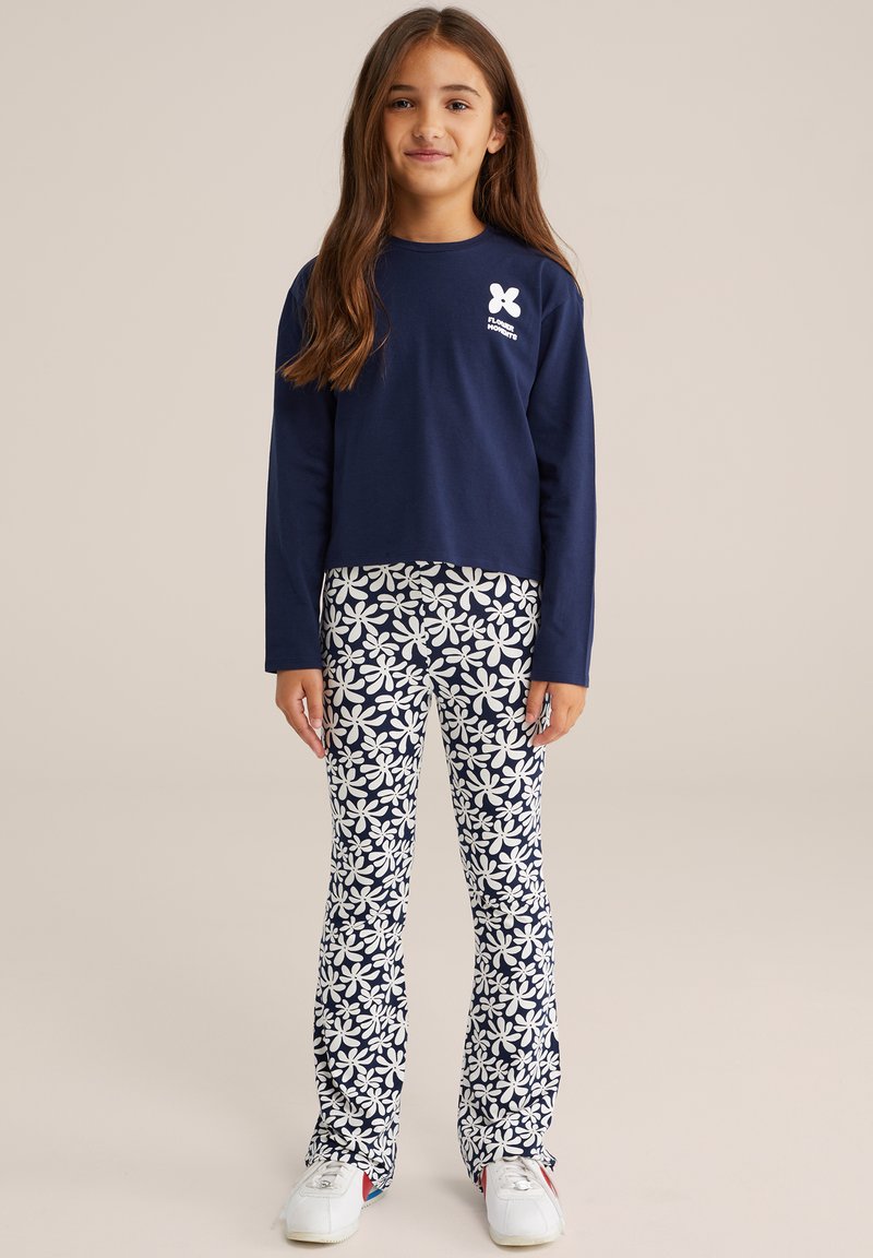 Mornarska majica z dolgimi rokavi z belim logotipom, v kombinaciji z belimi hlačami s cvetličnim vzorcem na dekletu, ki nosi bele športne copate z rdečimi detajli.