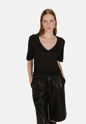 Femme aux cheveux rouges raides portant un haut noir à col en V à manches courtes et un short en cuir noir ample de longueur genou, les mains dans les poches.