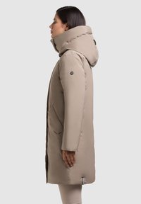 Béžový vatovaný kabát po kolená s vysokým golierom a kapucňou. Má bočné vrecká a hladký, lesklý povrch. Čierny logotyp na rukáve.