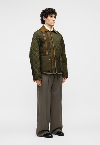 Άνδρας στέκεται φορώντας πράσινο καπιτονέ μπουφάν με καρό επιθέματα, φαρδιά καφέ παντελόνια και μαύρα παπούτσια μπροστά σε ένα απλό φωτεινό φόντο.