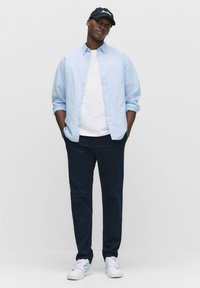 Helles blaues Button-up-Hemd über einem weißen T-Shirt, kombiniert mit dunklen Hosen und weißen Sneakers. Dazu eine marineblaue Baseballkappe mit weißem Text.