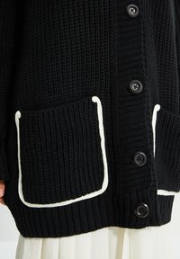 Svart stickad cardigan med ribbat mönster, vit kant runt fickorna och stora svarta knappar. Synliga fickor framtill.