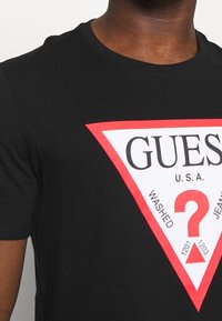Detailní záběr na osobu oblečenou v černém tričku s bílým trojúhelníkem s červeným okrajem a logem "GUESS U.S.A." se červeným otazníkem.