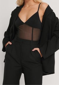 Blazer oversize nero sopra un body in rete nero, abbinato a pantaloni a vita alta neri e sartoriali. Caratteristiche includono spalline sottili e silhouette strutturata.