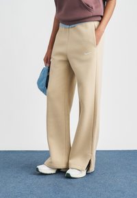 Μπεζ φαρδιά φόρμα με πλαϊνές σχισμές, διακοσμημένη με μικρό λευκό λογότυπο της Nike. Συνδυάζεται με λευκά αθλητικά παπούτσια και μπλε καπέλο από τζιν.