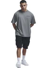 Homme portant un t-shirt gris oversize avec un logo, un short cargo noir, des chaussettes blanches et des baskets blanches, debout avec les mains dans le dos.
