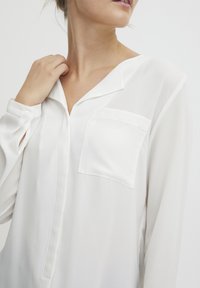 Blusa bianca a maniche lunghe con una leggera lucentezza. Presenta un colletto e una tasca frontale, con una vestibilità rilassata e un tessuto morbido e leggero.