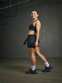 Femme en forme vêtue de vêtements de sport noirs et de baskets marchant à l’intérieur contre un mur sombre uni, souriante et regardant sur le côté.