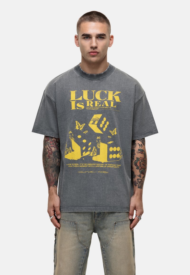 Szary, oversize T-shirt z żółtym napisem i graficznym wzorem przedstawiającym kości i motyle. Materiał wydaje się miękki, z lekko zużytą teksturą.