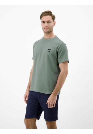 Man met lichtgroen gestructureerd shirt met korte mouwen en marineblauwe opgerolde korte broek, staand tegen een effen witte achtergrond.