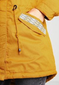 Žlutá bunda s texturovaným povrchem, která má viditelnou kapsu se vzorovaným akcentem a stahovací šňůrku v dolním lemu.