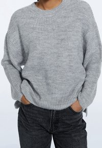 Gray knitted sweater with a round neckline and ribbed texture, featuring dropped shoulders and a relaxed fit, paired with dark jeans.