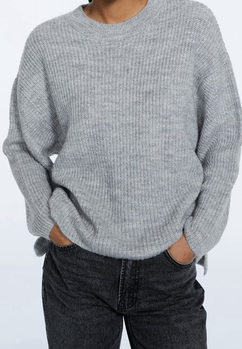 Gray knitted sweater with a round neckline and ribbed texture, featuring dropped shoulders and a relaxed fit, paired with dark jeans.