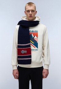 Marineblå ribbede halstørklæde med røde og hvide detaljer, der har et norsk flag lap. Bæres over en cremefarvet hoodie og sat sammen med sorte bukser.