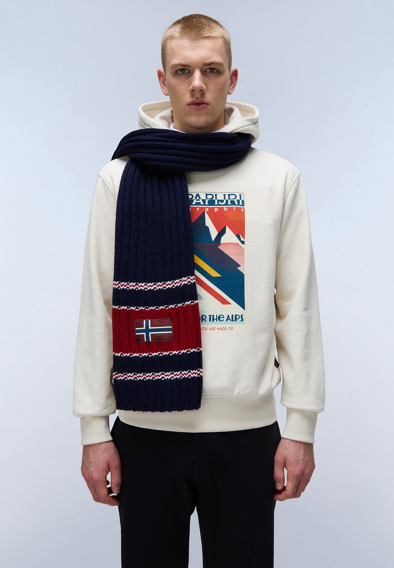 Marineblå ribbede halstørklæde med røde og hvide detaljer, der har et norsk flag lap. Bæres over en cremefarvet hoodie og sat sammen med sorte bukser.