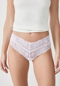 Intimo in pizzo bianco con motivi floreali, bordi ondulati e una texture semitrasparente. Abbinato a una maglietta bianca aderente.