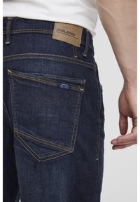Jeans de mezclilla en azul oscuro con costuras amarillas en contraste. Presentan un parche de marca de cuero en la cintura y un bolsillo inferior con una etiqueta.