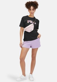 Camiseta gráfica negra con el logo "FILA", pantalones cortos lavanda y riñonera rosa. Zapatillas blancas completan el conjunto casual.
