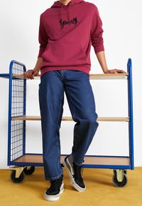 Felpa bordo con logo nero, abbinata a jeans blu larghi e sneakers nere con accenti bianchi e grigi, appoggiato a un carrello blu.