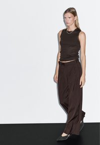 Brązowy top na ramiączkach, luźne brązowe spodnie i ciemnoszare buty. Gładki materiał, dopasowany top i detal z guzikami na spodniach. Minimalistyczny design.