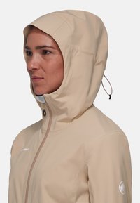 Beige wasserdichte Jacke mit angepasster Kapuze, kontrastierendem Reißverschluss und elastischen Anpassungen. Verfügt über ein Logo auf dem Ärmel und eine glatte Textur.