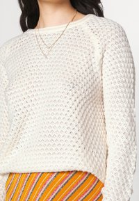 Pull tricot blanc avec un motif texturé et un col rond, associé à une jupe rayée multicolore avec des accents orange et rose.